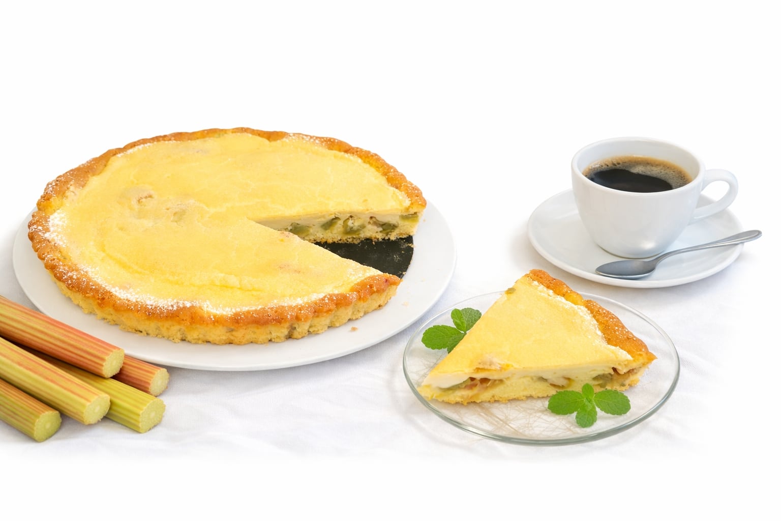 cremiger Rhabaraberkuchen angeschnitten auf einem weißen Tortenteller, Rhabarberstangen als Deko und ein Stück serviert auf einem Glasteller, im Hintergrund eine Tasse Kaffee