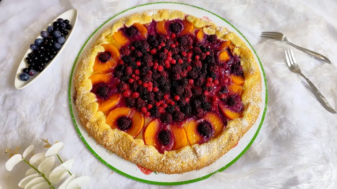Rustikale Galette mit frischen Früchten wie Pfirsichen und Beeren, goldbraun gebacken und auf einem Tortenteller serviert - ein köstliches hausgemachtes Dessert