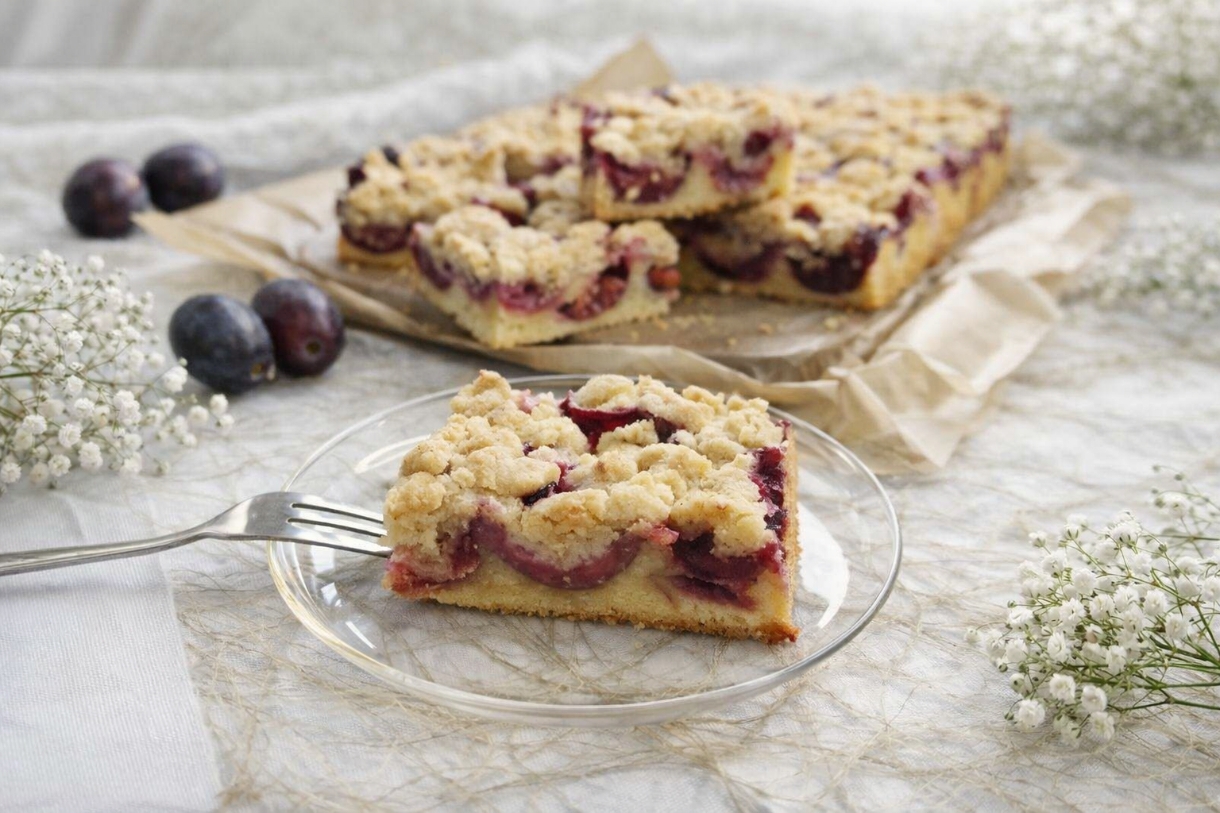Frisch gebackener Pflaumenkuchen mit Rührteig und goldbraunen Streuseln, belegt mit saftigen Pflaumenhälften - serviert auf dem Backblech mit Backpapier, ein Stück serviert auf einem Glasteller im Vordergrund