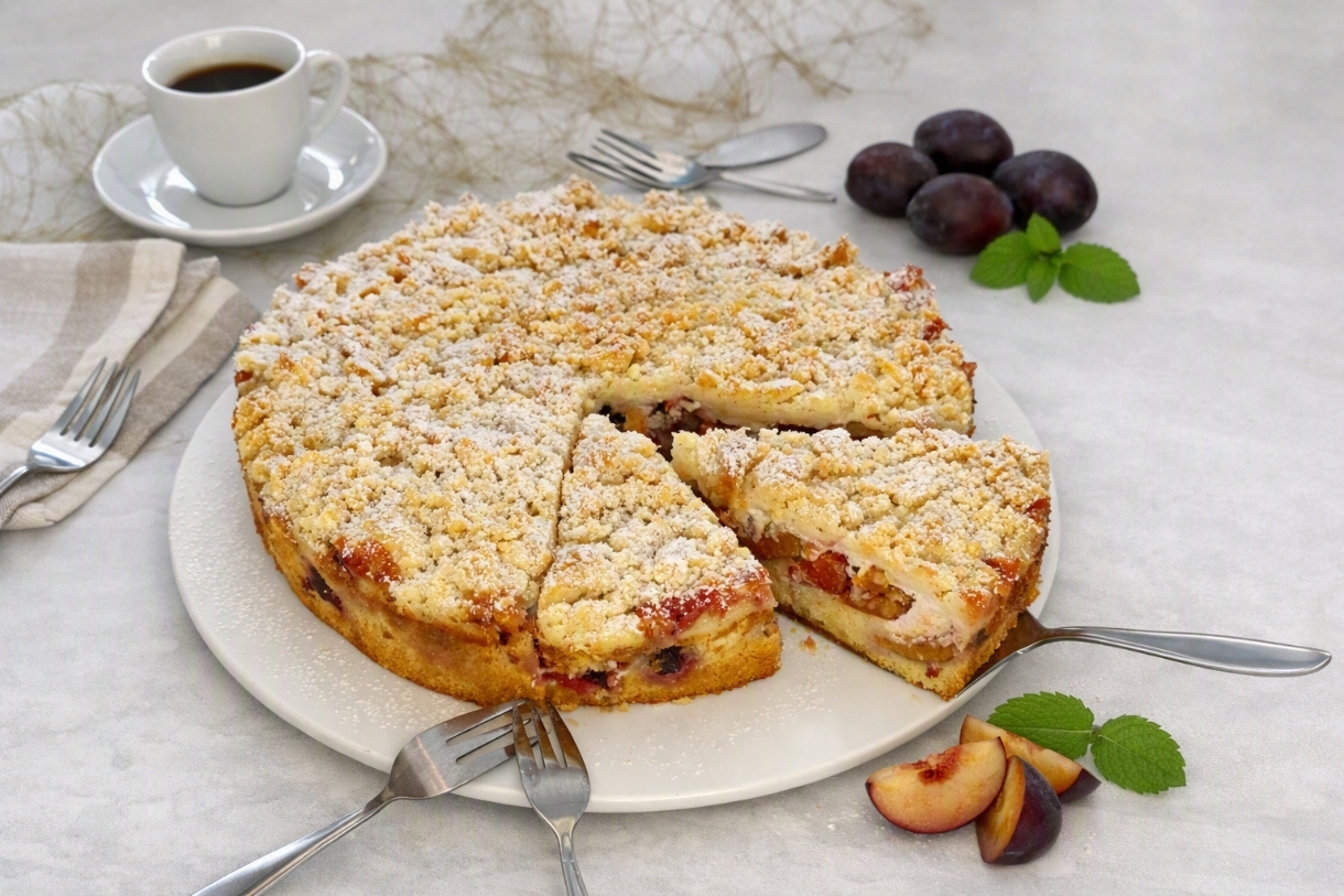 Saftiger Pflaumenkuchen mit Streusel gebacken in einer Springform, angeschnitten, im Hintergrund eine Tasse Kaffee und Pflaumen als Dekoration