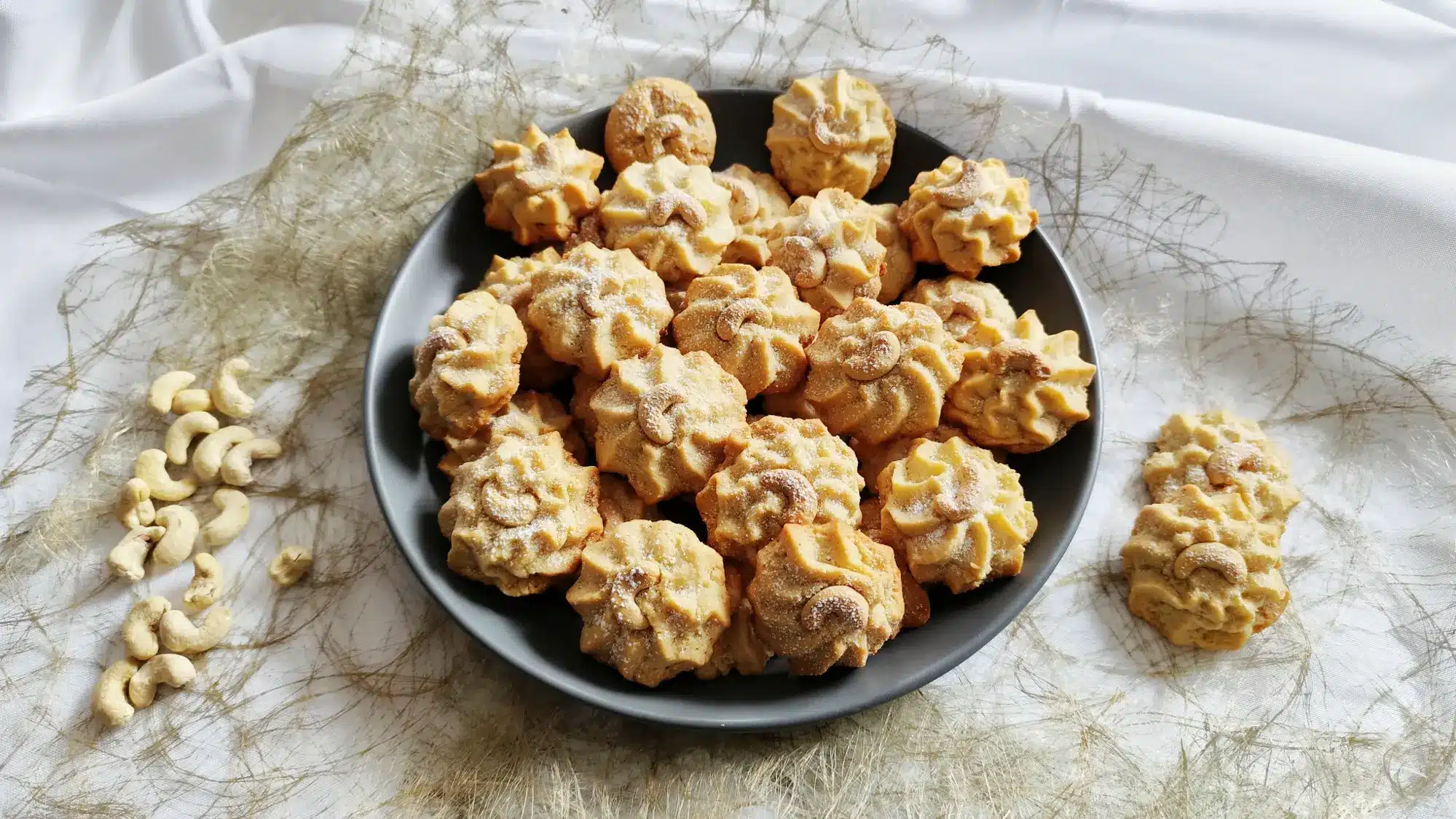 Goldgelbene Cashew-Plätzchen auf einem dunklen Keramikteller, dekoriert mit Cashewkernen