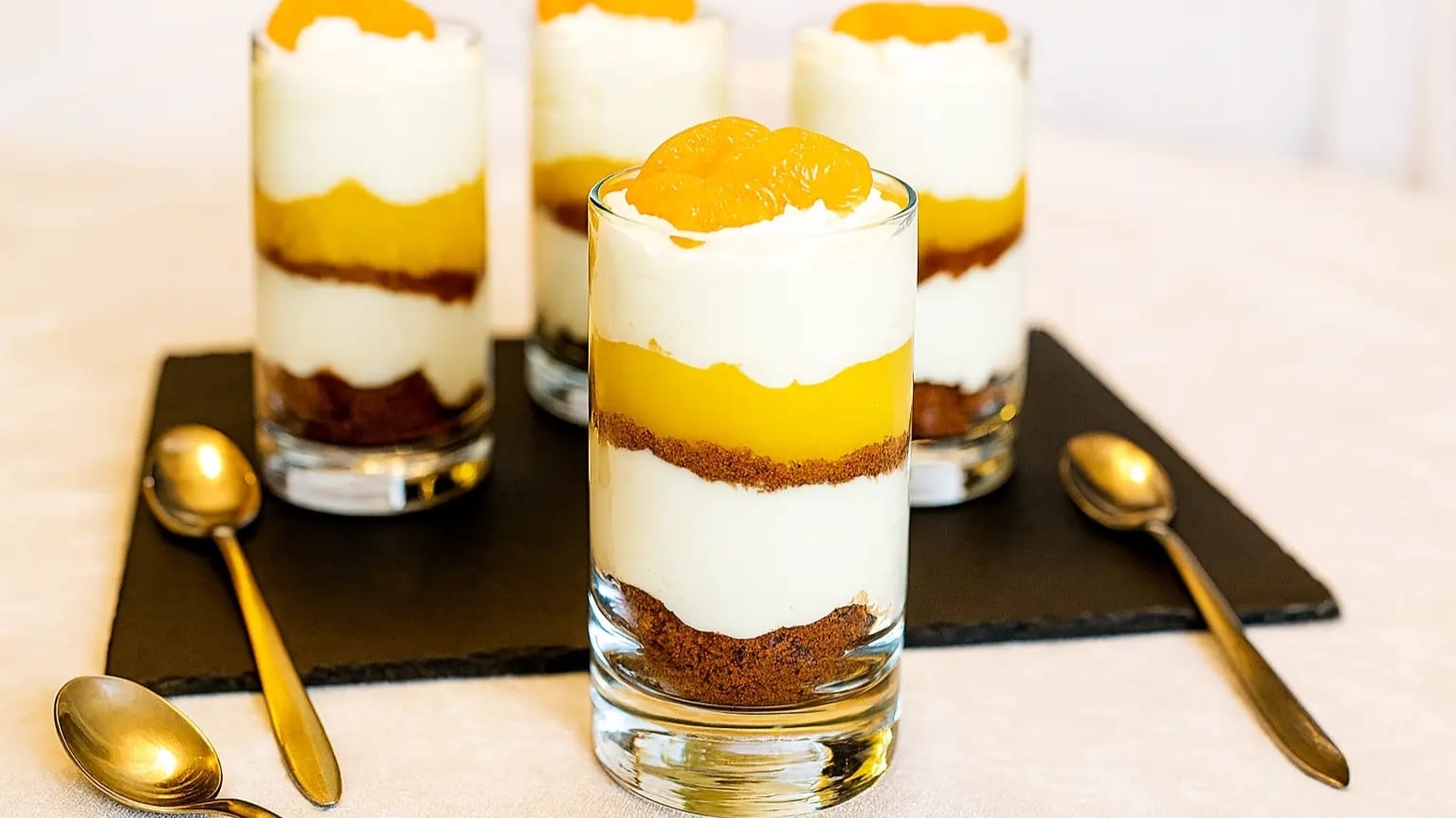 Fruchtiges Schichtdessert im Glas mit goldenen Löffeln auf schwarzem Servierbrett