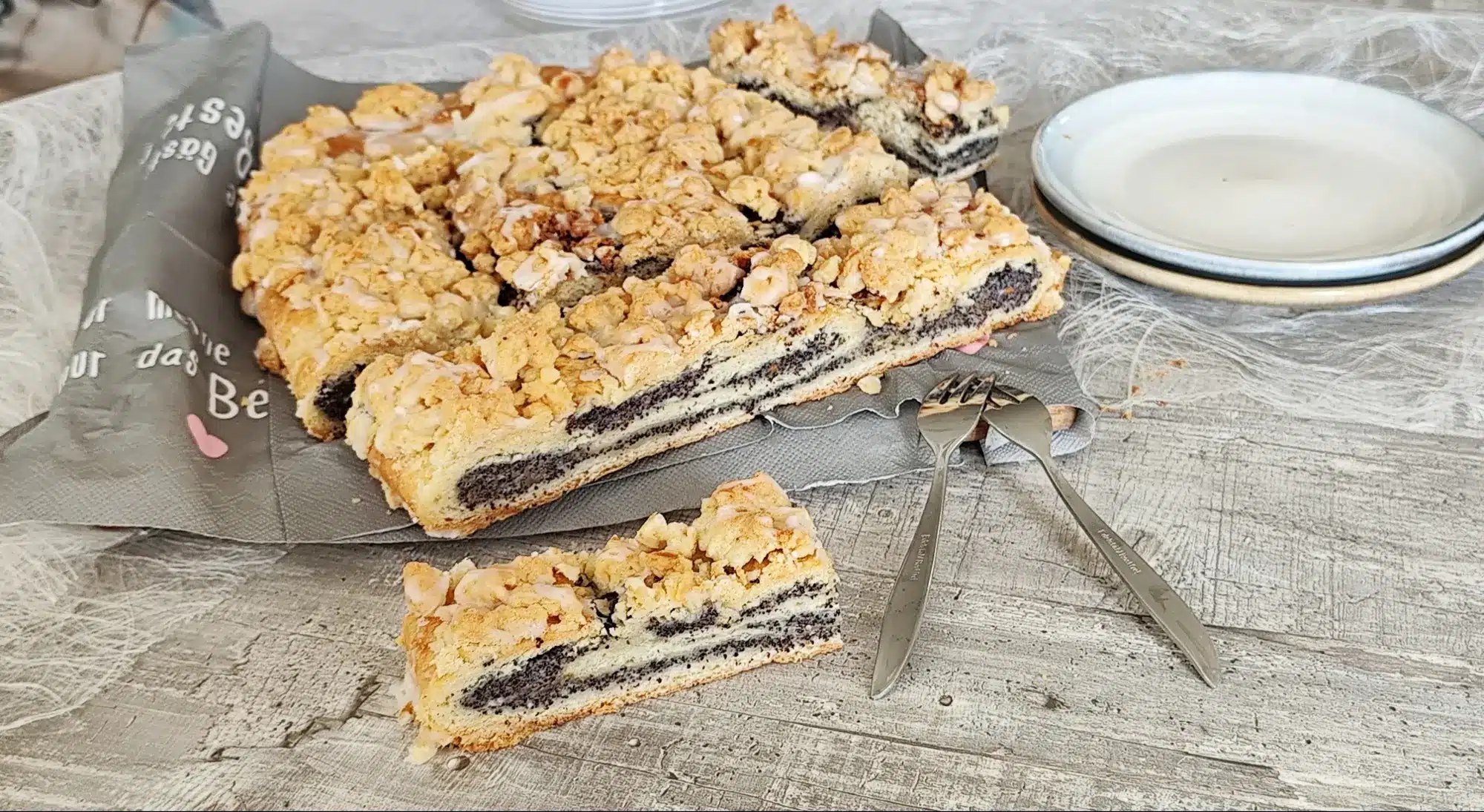 Saftiger Mohnstriezel vom Blech, in Stücke geschnitten, mit dicker Mohnfüllung und knusprigen Streuseln, mit Tellern und Kuchengabeln im Hintergrund