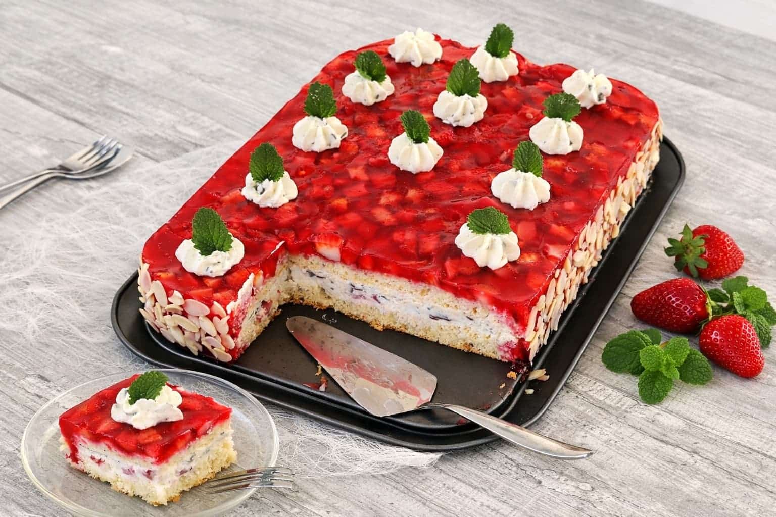 Erdbeerschnitten mit Stracciatellacreme und Sahnetuffs mit Minzblättern auf einem Blech; ein Stück auf einem Glasteller angerichtet, dekoriert mit frischen Erdbeeren und Mandeln