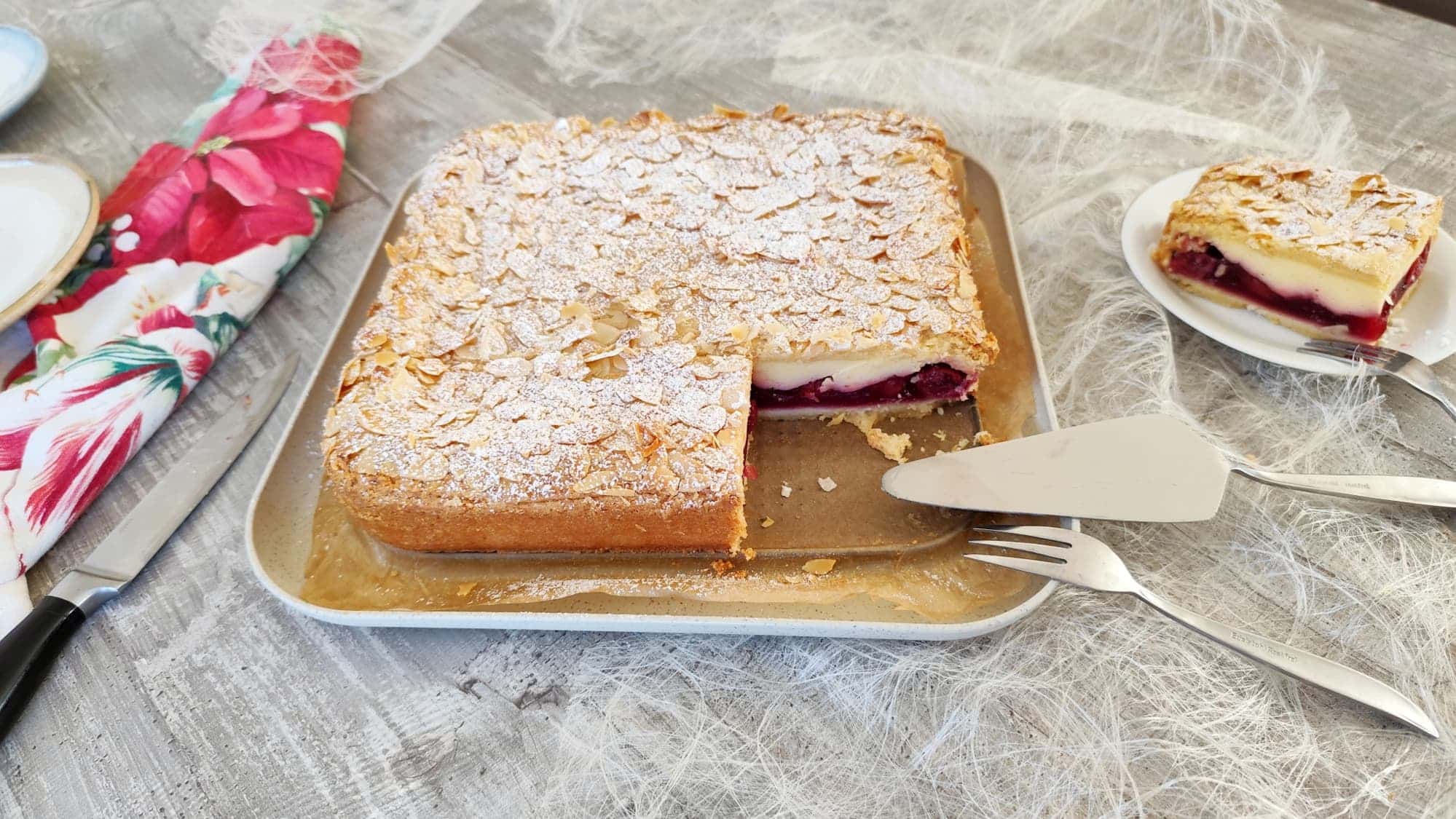 Gedeckter Kirschkuchen mit Schmad-Quarkfüllung und goldbrauner Mandeldecke auf einem hellen Teller, angeschnitten mit Kuchengabel und Tortenheber, seitlich ein Stück auf einem weißen Teller