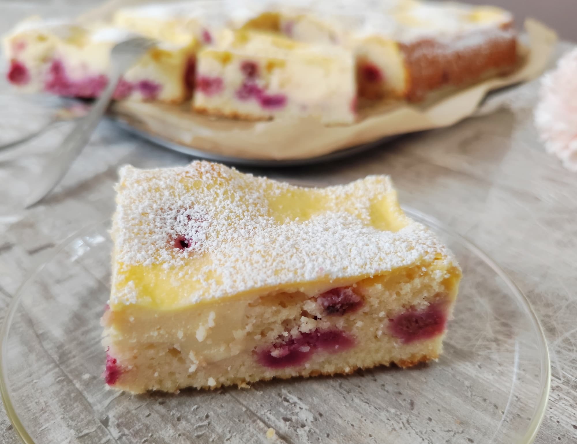 Ein Stück Buttermilchkuchen mit Kirschen und Quarkguss, dekoriert mit Puderzucker, serviert auf Glasteller, im Hintergrund angeschnittener Kuchen auf Backblech